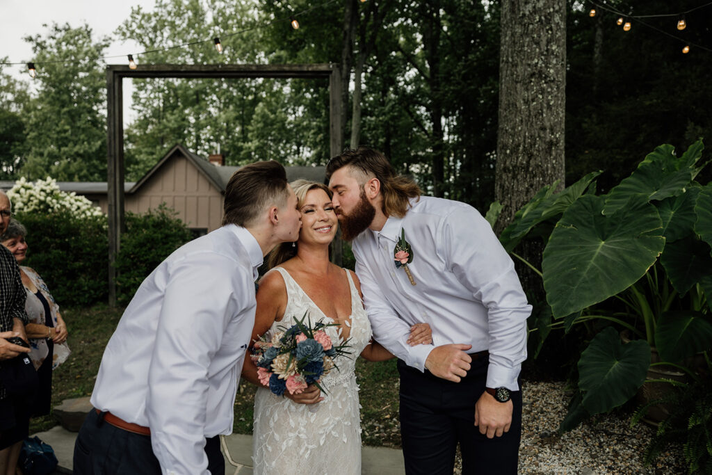 A bride is walked down the aisle by her two sons. They are both leaning in and kissing her cheek. She is smiling