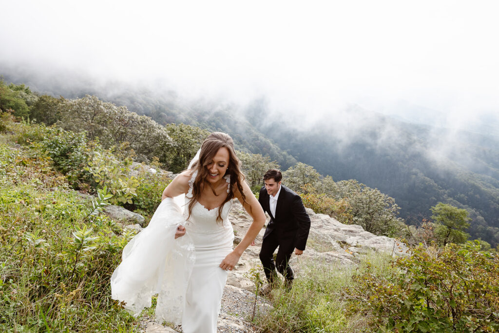 a bride and groom hike up a hill. You can see the mountains and fog behind them. The bride is holding up her veil and dress. The groom is wearing a black suit