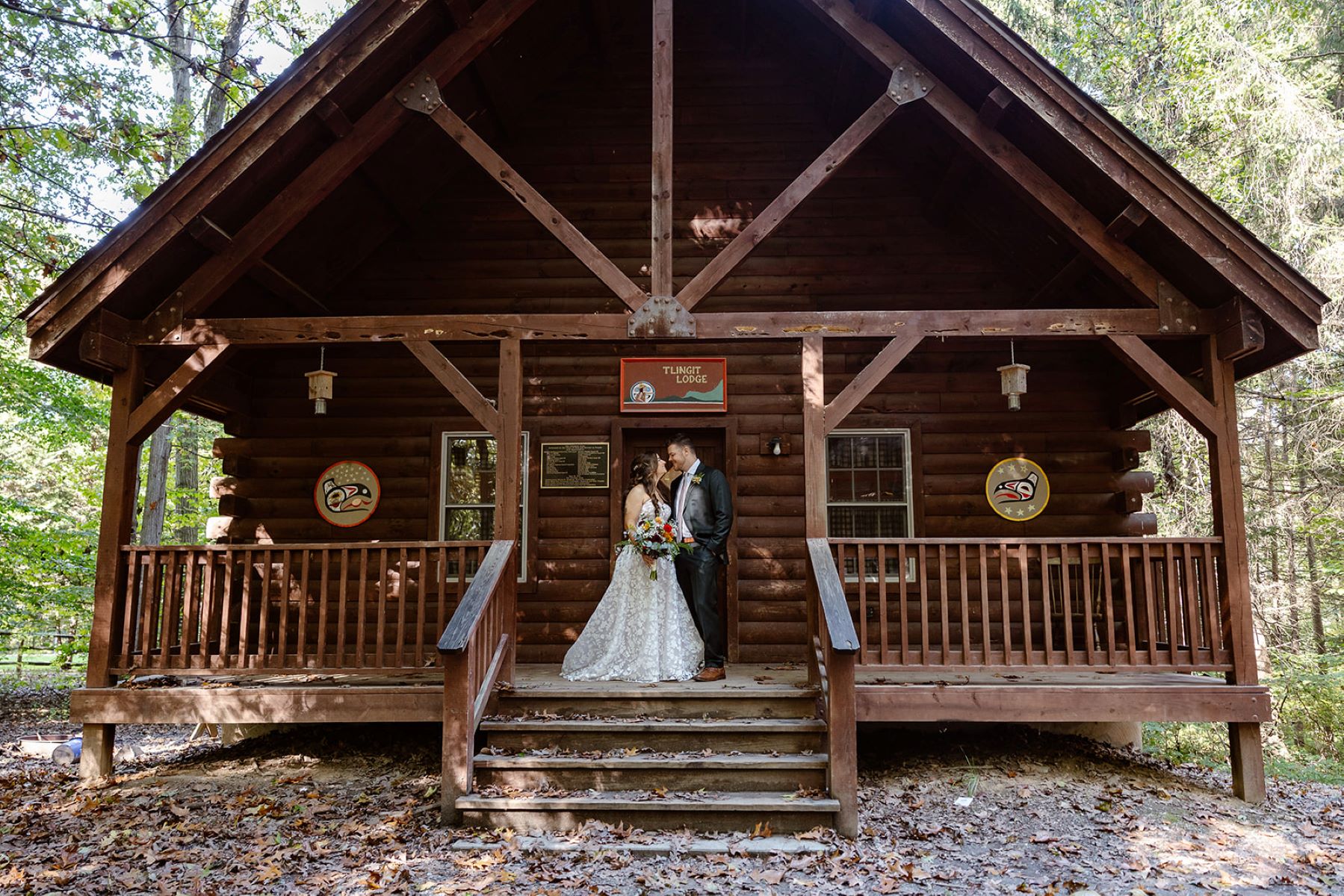 A woman in a white wedding dress holding a bouquet of flowers and a man in a black suit standing on the porch of a wooden lodge and smiling at each other 