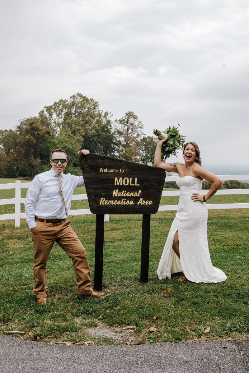 A woman in a white wedding dress is laughing and holding her bouquet in the air and a man in a white shirt and tan pants who is also  wearing sunglasses is smiling and they are both standing next to a wooden sign 