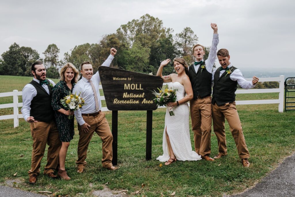 A woman in a white wedding dress with her partner with their wedding party standing next to a wooden sign on a patch of green grass celebrating 