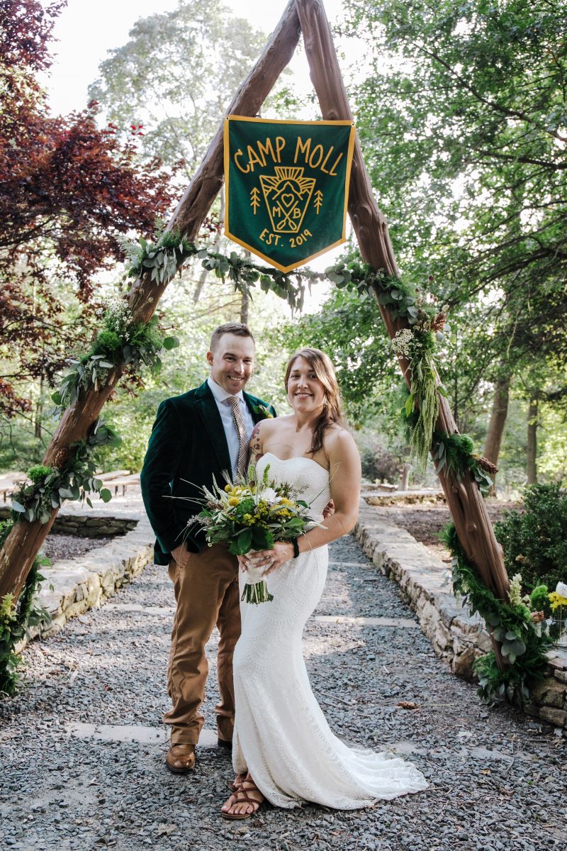 A woman in a white wedding dress is holding a bouquet of flowers and smiling and a man in a blazer and tan pants is smiling and standing next to her and behind them is a wooden arch