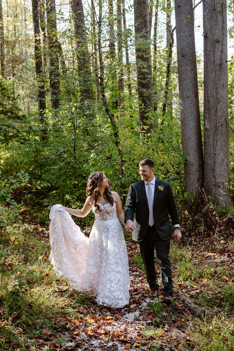 A woman in a white wedding dress and a man in a black suit and walking through the woods holding hands and smiling at each other 