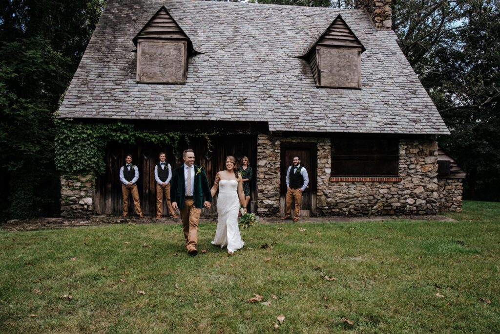 A couple in front of a lodge the woman is wearing a white dress the man is wearing a white dress jacket and tan pants and they are walking together on a lawn and their wedding party is behind them watching them