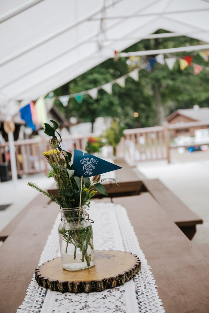 Table decor for a campground wedding. There are custom flags in the flower vases. There are long picnic tables with lace table runners