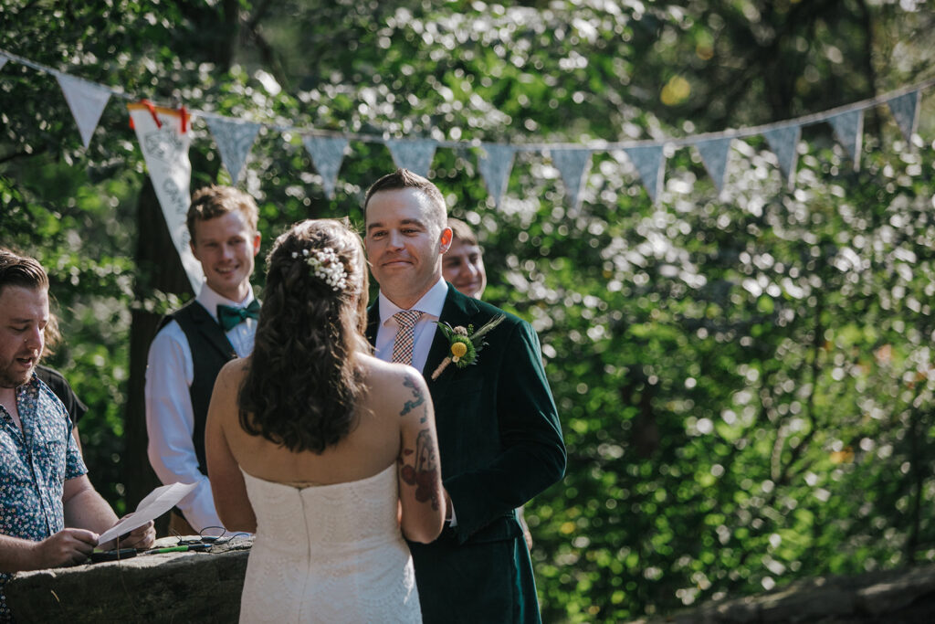 A bride and groom during the vows of their wedding ceremony. The bride is wearing a strapless dress, and the groom is wearing a green velvet jacket. There is a pennant banner hanging above them.