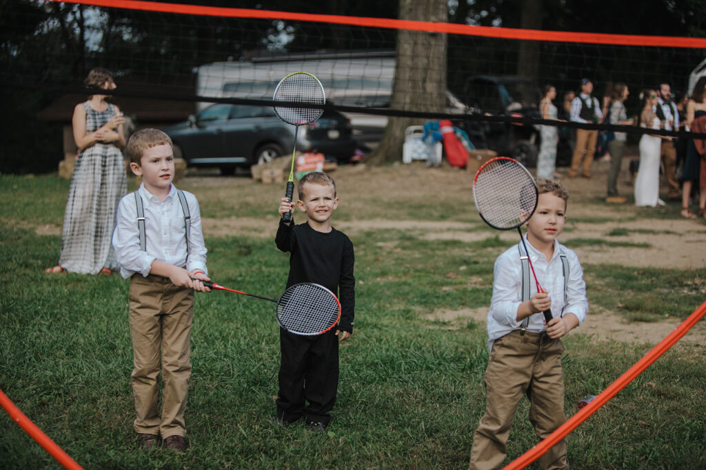 Three children stand holding up badminton rackets. Two are dressed in white shirts, brown pants, and green suspenders. 