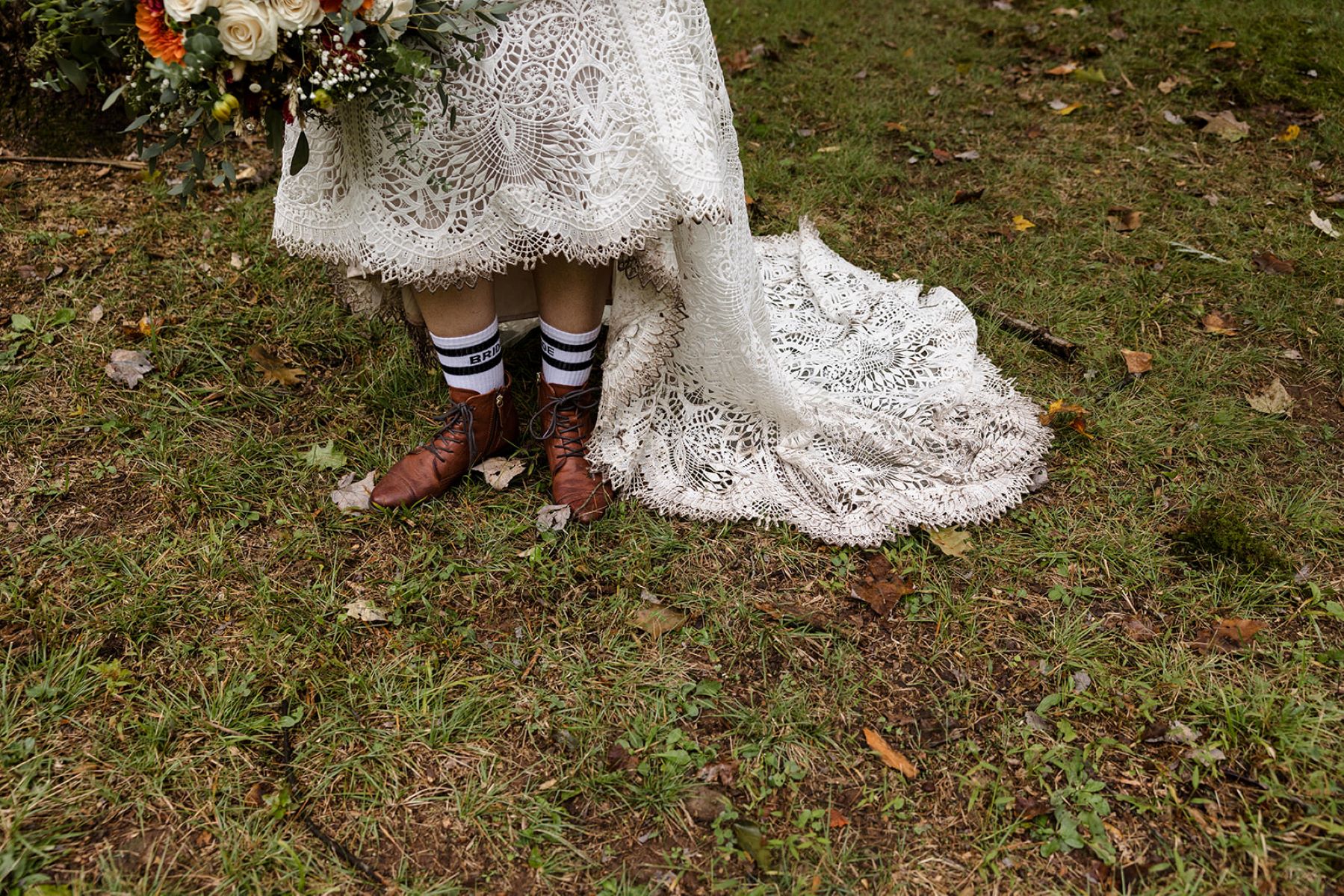 A woman wearing a white wedding dress standing in the grass wearing hiking boots and holding a bouquet of flowers 