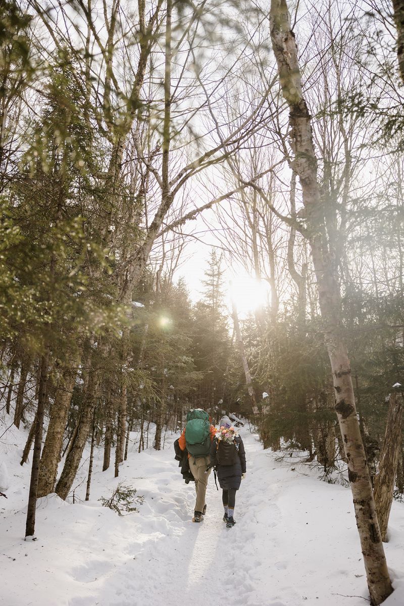 A couple walking through a forest and the snow wearing backpacks and winter attire 