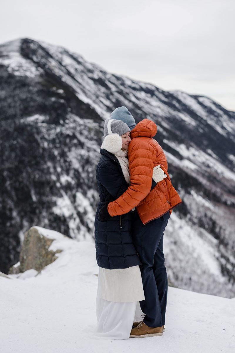 A couple on a mountain top covered in snow hugging each other they are both wearing winter attire 