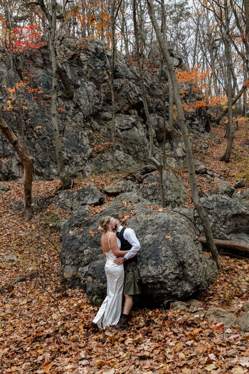 A couple standing next to a large rock and standing on top of leaves and around them are large boulders and a forest the woman is wearing a white wedding dress the man is wearing a kilt and kissing his partner 