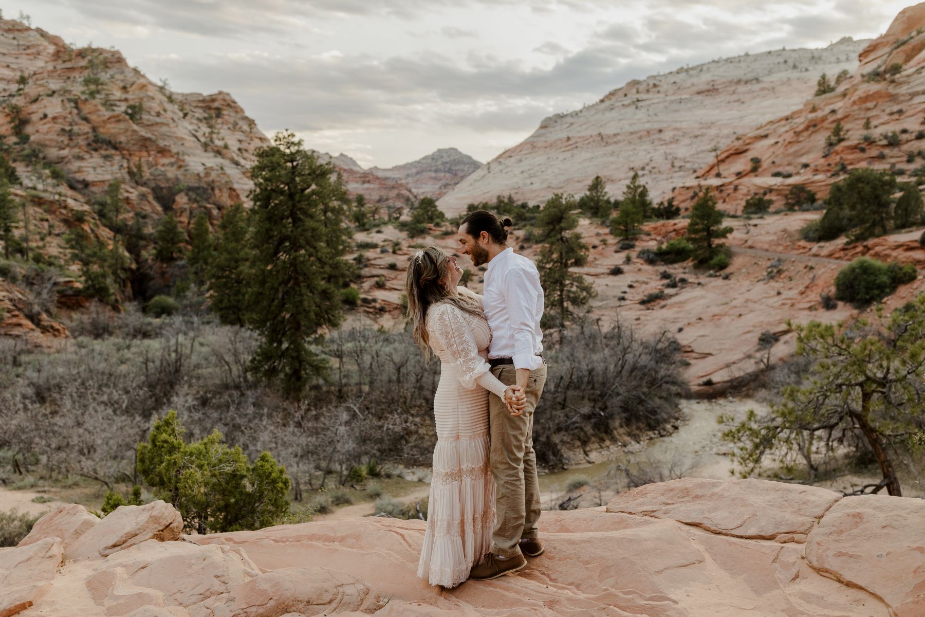 A couple standing on top of a cliff with a valley behind them they are holding hands and smiling at each other the woman is wearing a dress and the man is wearing awhite button up and tan pants