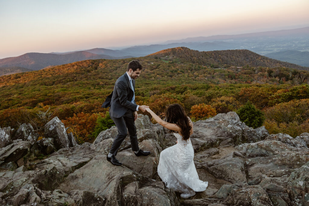 An eloping bride and groom climb to the top of a lookout in Shenandoah National Park. The groom is helping the bride in a romantic way.