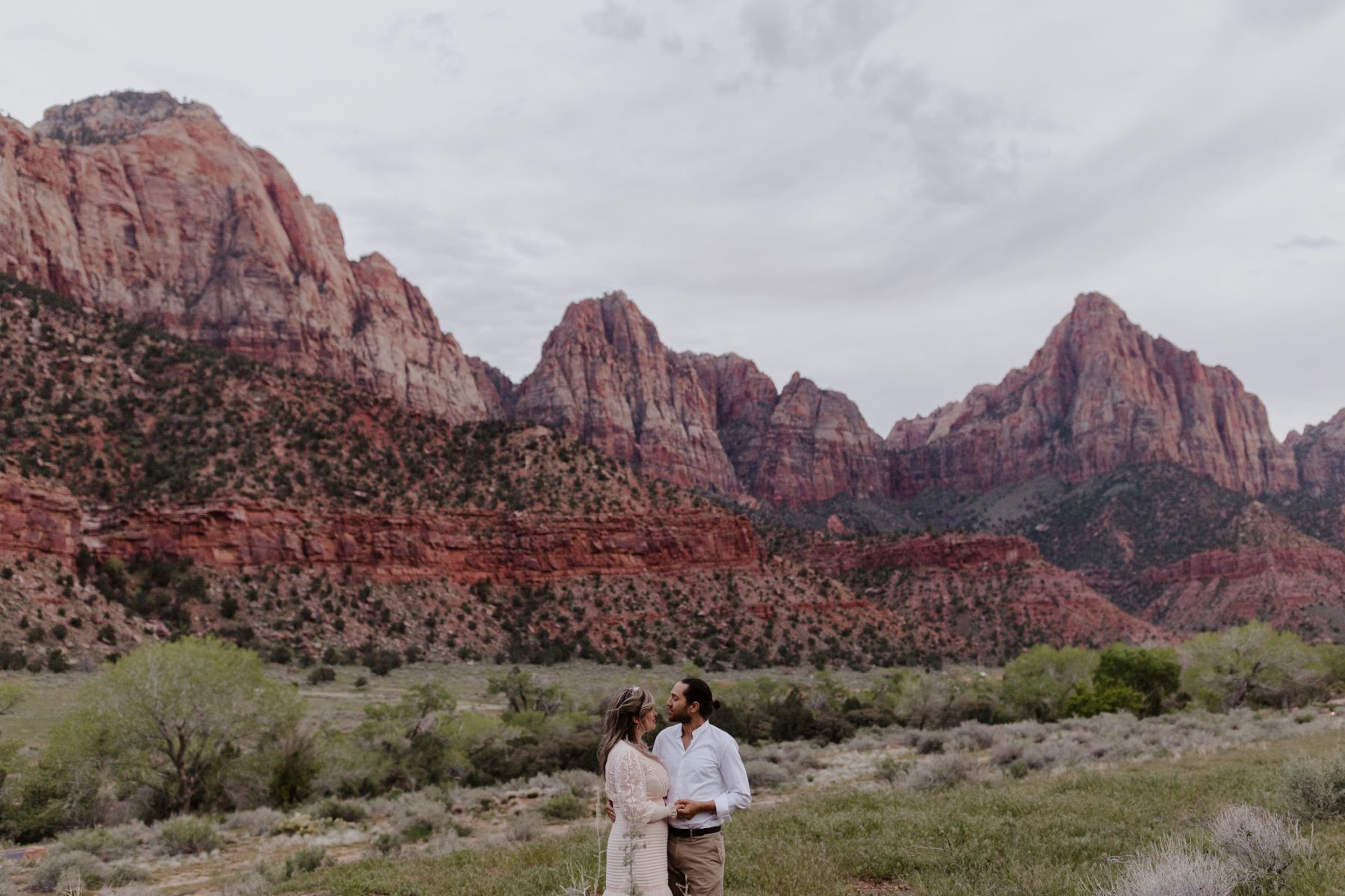 A couple standing in a valley that is surrounded by mountains they are looking into each other's eyes and holding hands the woman is wearing a white wedding dress and the man is wearing a white button up and tan pants