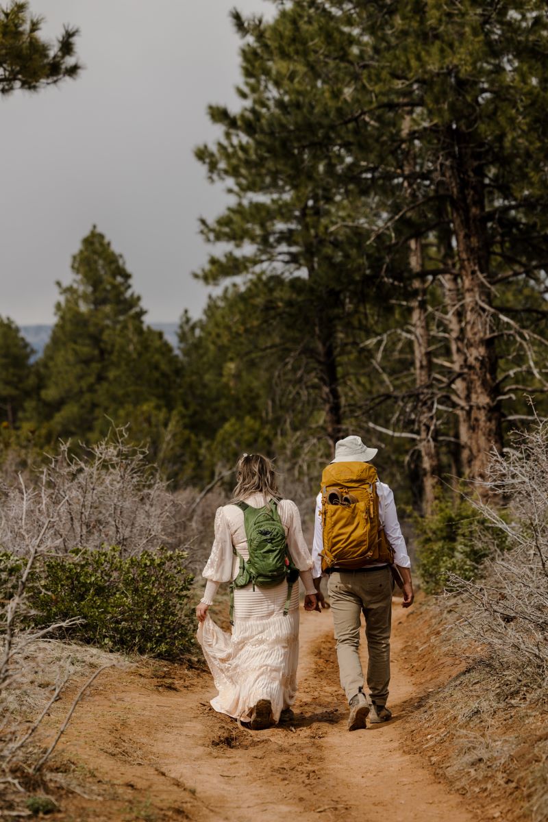 A couple walking on a trail together with their backpacks on and holding hands the woman is wearing a white wedding dress and has a green backpack the man is wearing a hat, a button up and pants and has a yellow backpack 