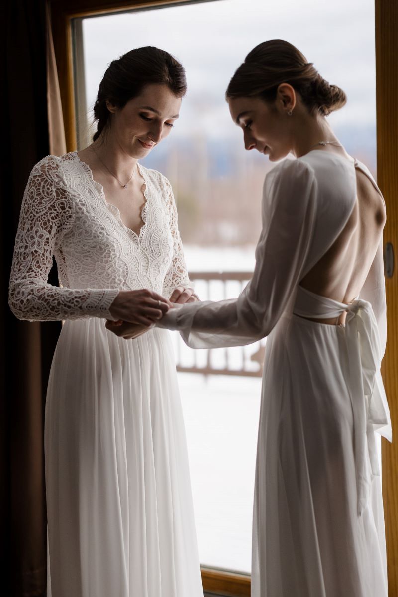 Two women standing together wearing white wedding dresses one woman is helping the other woman adjust her dress sleeve