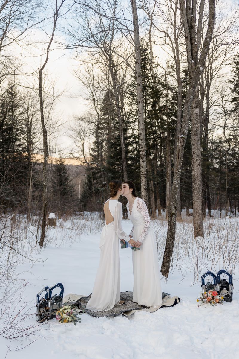 Two women at their wedding ceremony kissing and touching hands they are outdoors and the ground is covered by snow 