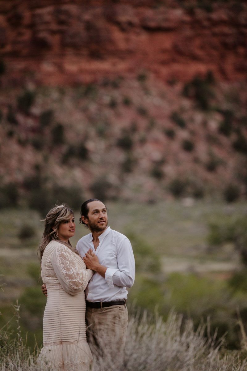 A couple standing in a field looking into the distance with their hands holding each other the woman is wearing a white wedding dress the man is weairng a white button up and khaki pants