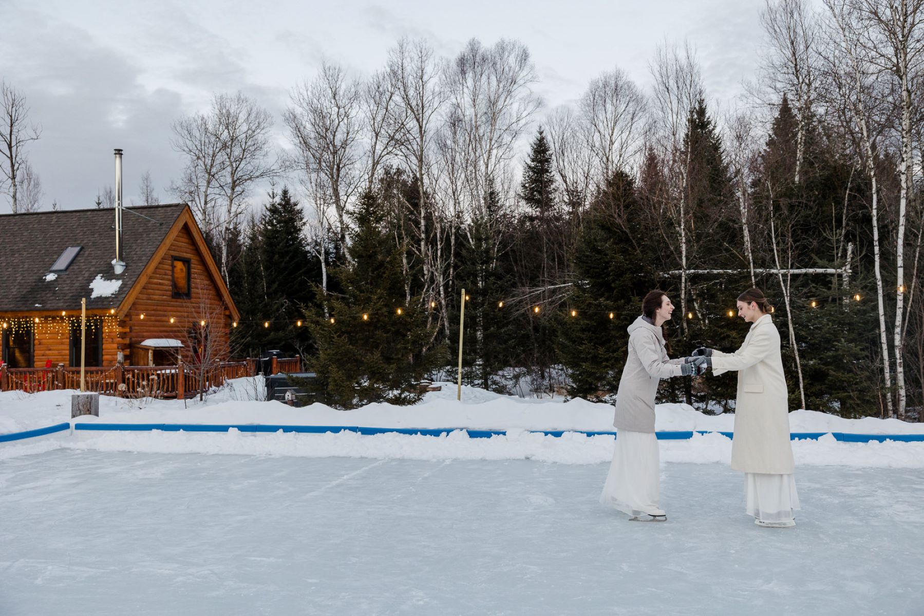 Two women on an ice skating rink holding hands and laughing they are both wearing white wedding dresses and coats 