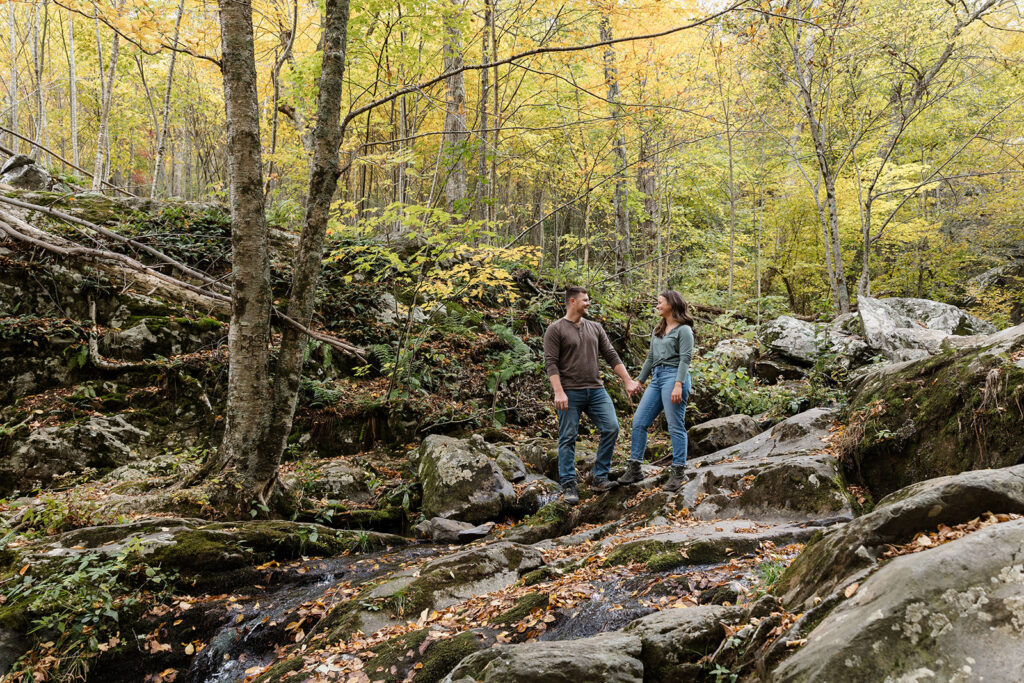 A couple holds hands during their Shenandoah Adventure Session in fall. They are standing in a boulder field near Dark Hollow Falls. they are wearing jeans and hiking boots. 