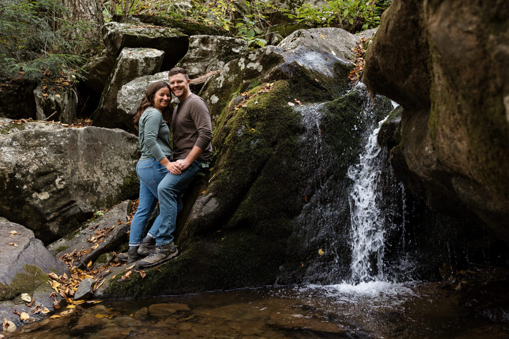 A woman lovingly leans into fiance during their Shenandoah Adventure Session in fall.  They are standing  holding hands next to Dark Hollow Falls. They are both wearing jeans and hiking boots