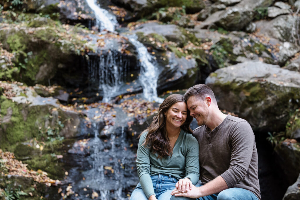 An engaged couple sitting on some boulders in front of Dark Hollow Falls in Shenandoah. They are holding hands, and leaning their foreheads toegther