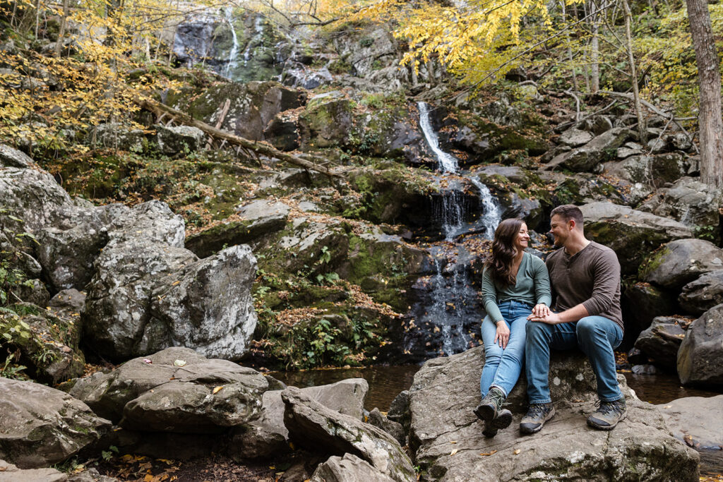 An engaged couple sitting on some boulders in front of Dark Hollow Falls in Shenandoah. They are holding hands, and and smiling at one another