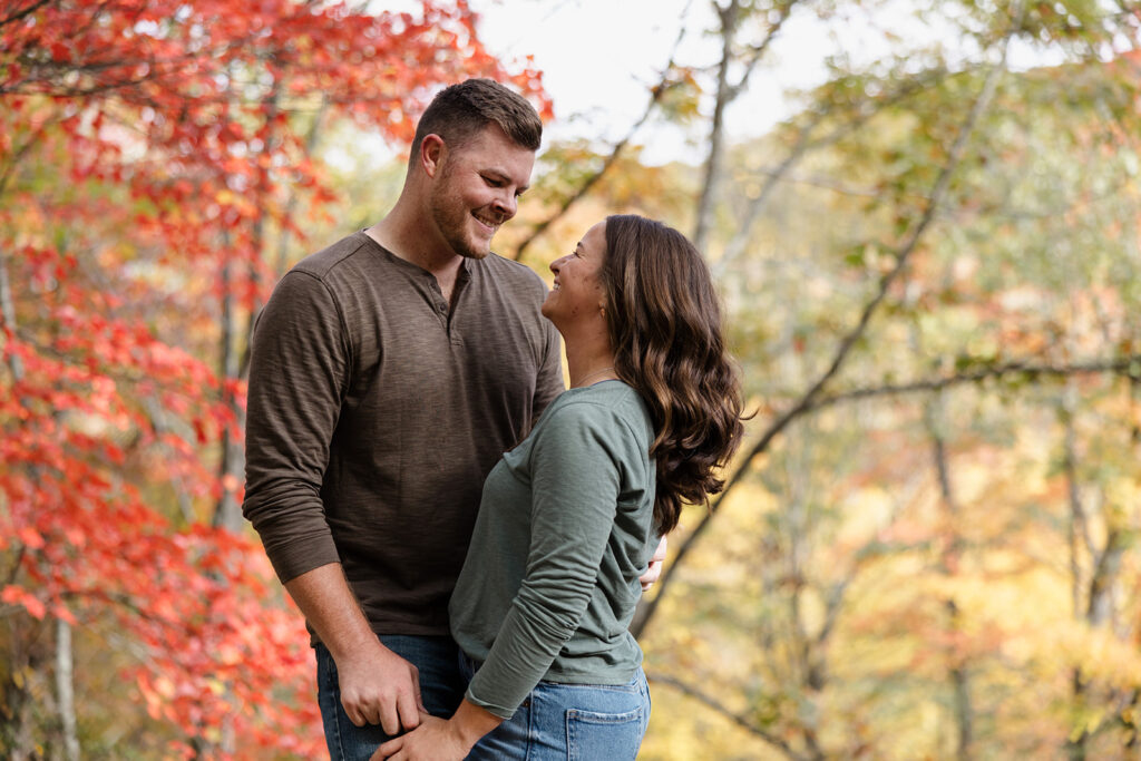 A couple stands in fall foliage in Shenandoah. They are facing each other  while smiling and holding hands. There are red leaves behind them