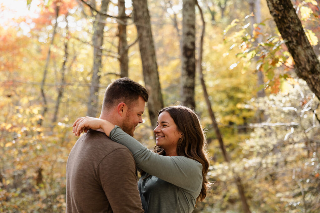 A couple stands in fall foliage in Shenandoah. They are facing each other  while smiling and holding hands. 