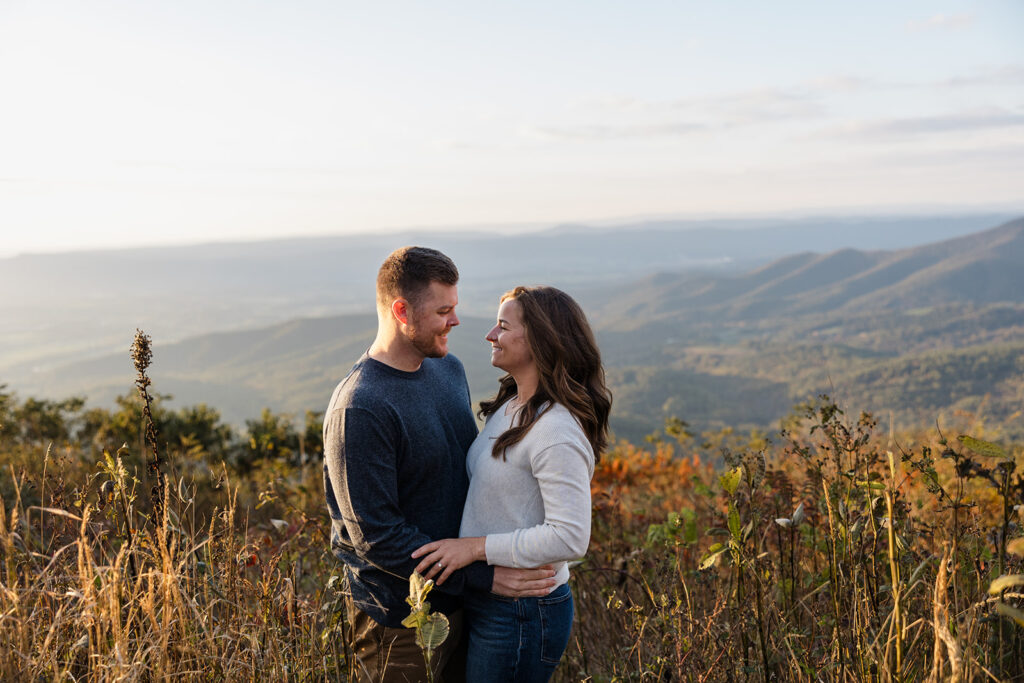 An engaged couple are standing in a field during their Shenandoah Adventure Session in fall. They are facing one another and smiling, You can see mountains behind them.