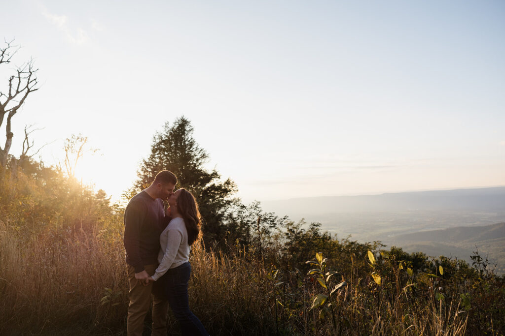 An engaged couple are standing in a field during their Shenandoah Adventure Session in fall. They are kissing as the sun sets behind them. You can see mountains in the background.