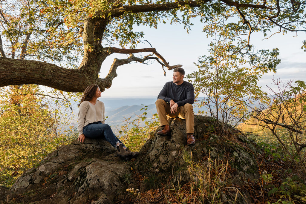 A couple sits on separate parts of a giant rock. You can see mountains in the background. They are looking at one another smiling. You can also see fall foliage in the background