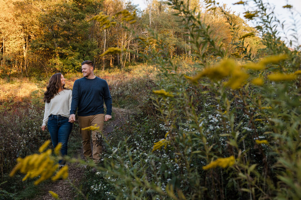 An engaged couple is holding hands and walking in a field of goldenrod during their Shenandoah Elopement Session in fall.