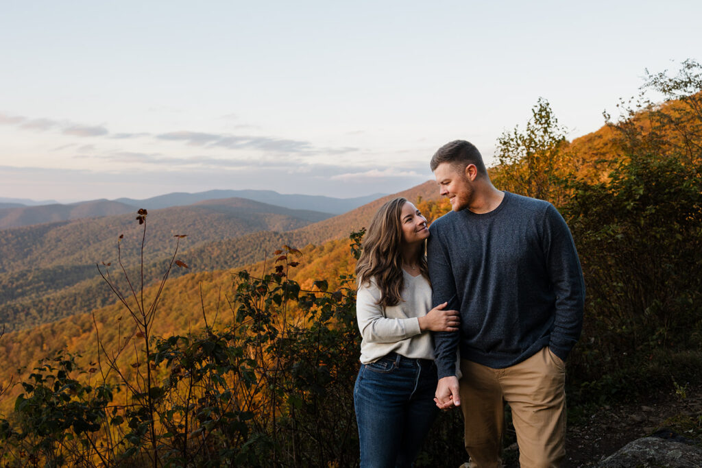 An engaged couple standing next to one another in front of some fall foliage during their Shenandoah Adventure Session in fall. They are looking at one another lovingly.