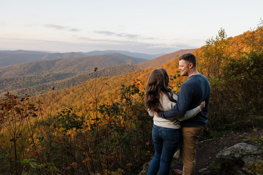 n engaged couple with their arms around one another in front of some fall foliage during their Shenandoah Adventure Session in fall. They are looking at one another lovingly.