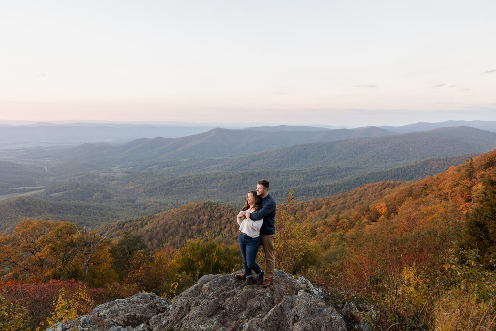 An engaged couple snuggling in front of. some fall foliage during their Shenandoah Adventure Session in fall. The man is standing behind the woman, and they are holding hands