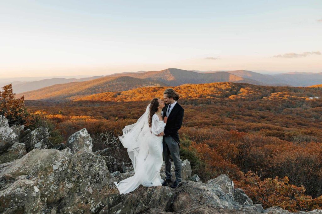 A bride and groom hold each other closely during their chilly and windy sunrise elopement at Shenandoah National Park. The foliage is near peak colors in the background