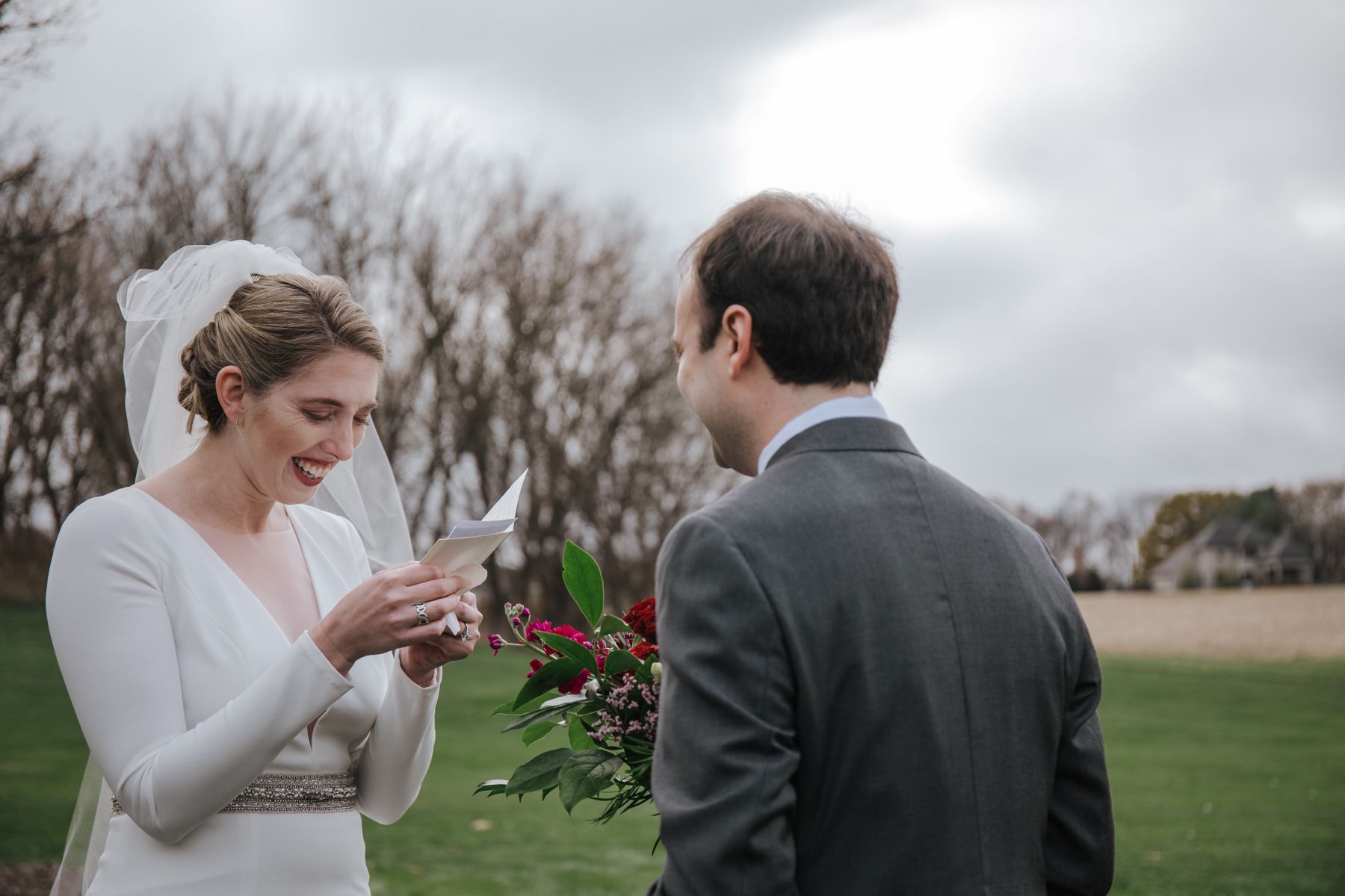 a woman holding a vow book and reading it while her partner stands in front of her and is looking at her the woman is wearing a veil and a white wedding dress 