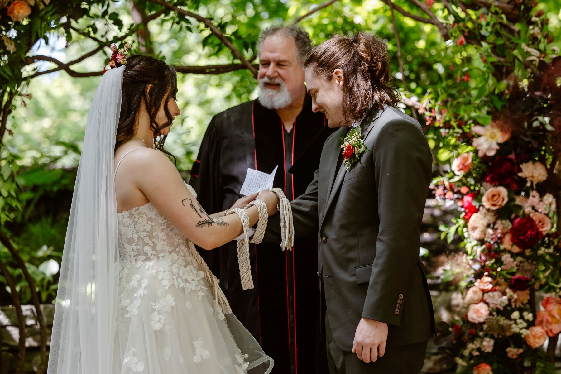 a man and a woman with their officiant performing a handfasting ceremony the woman is wearing a white wedding dress and veil the man is wearing a suit and a cord is wrapped around their hands their officiant is watching them 