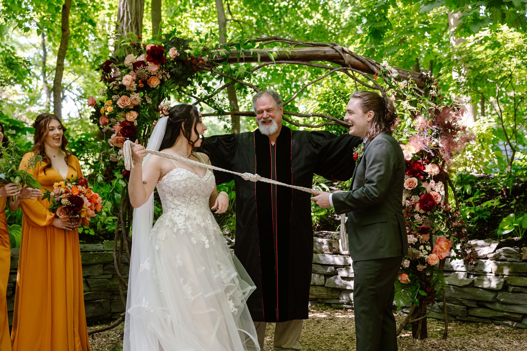 a man and a woman at their wedding ceremony performing a hand fasting ceremony the woman is tuggin the cord back and her partner is laughing and their offiicant is standing behind them smiling