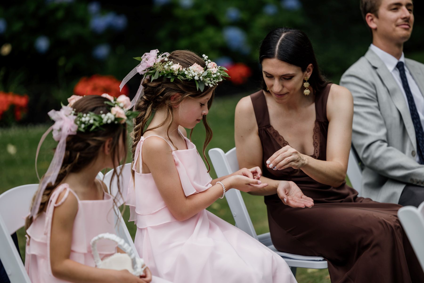 two girls wearing pink dresses and flower crowns and sitting in white chairs one girl is holding a wedding ring and a weding guest is sitting next to her and reaching for the ring