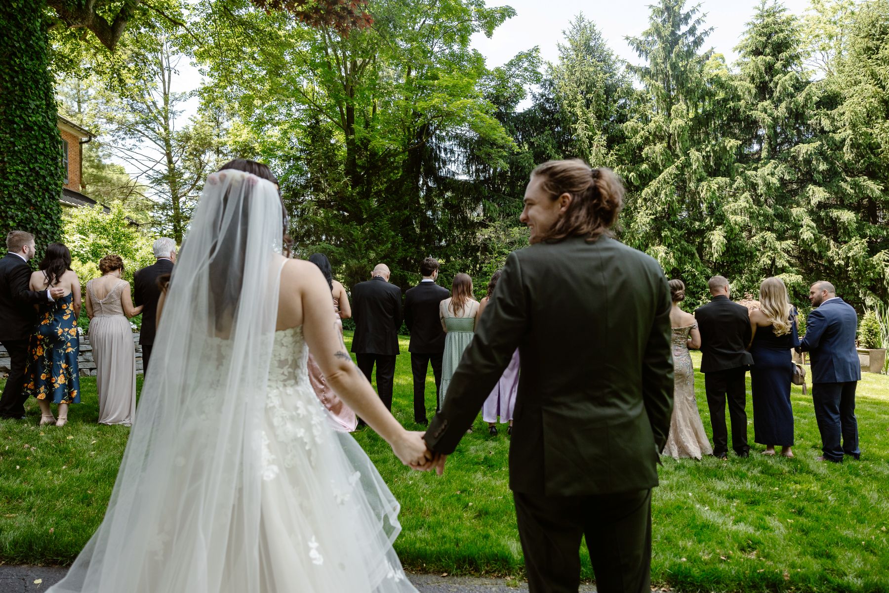 a couple holding hands and looking at their wedding guests who have their backs turned to them the woman is wearing a white veil and dress the man is wearing a suit 