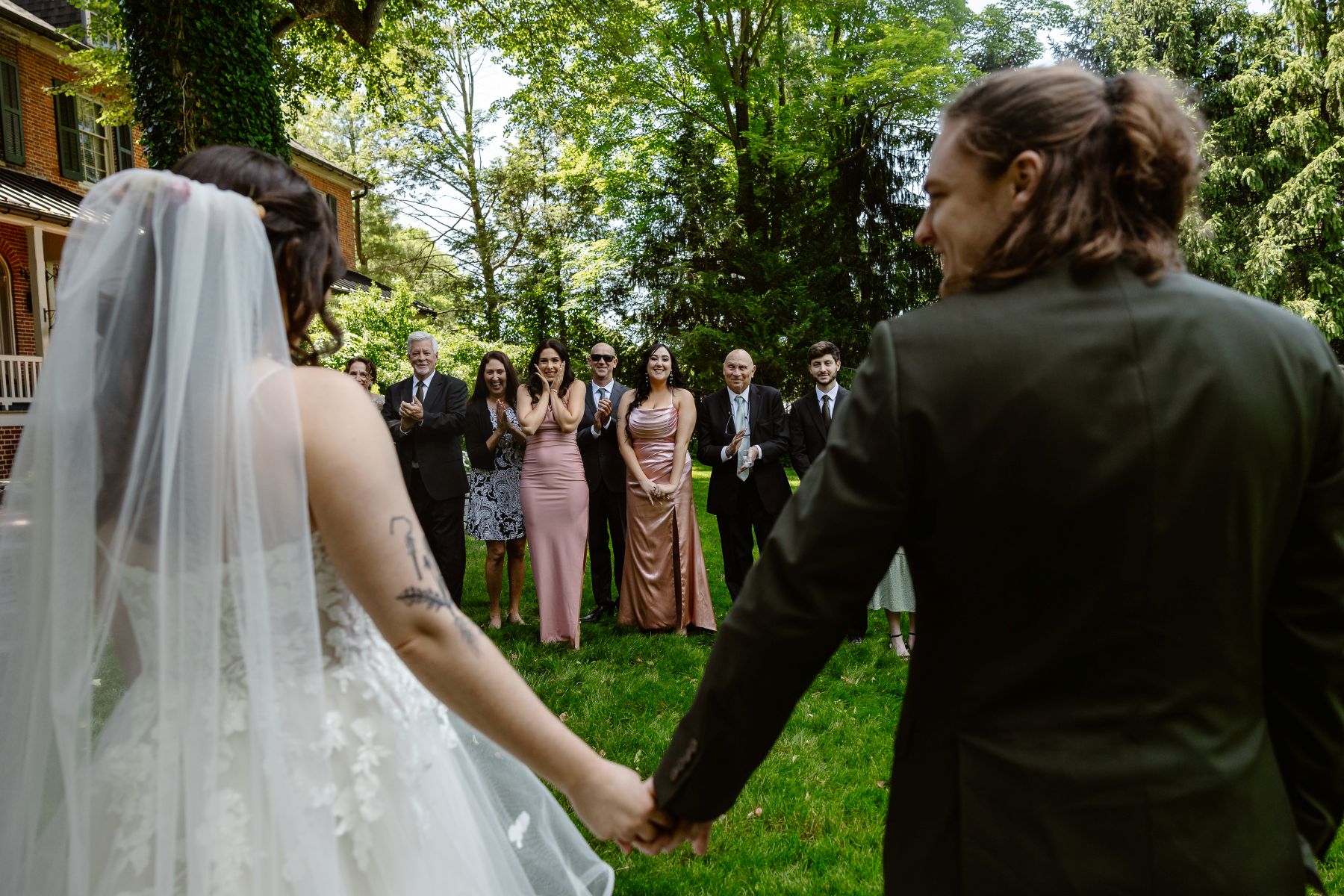 a couple doing a first look with their wedding guests the woman is wearing a white wedding dress and veil the man is wearing a suit and the couple is holding hands 