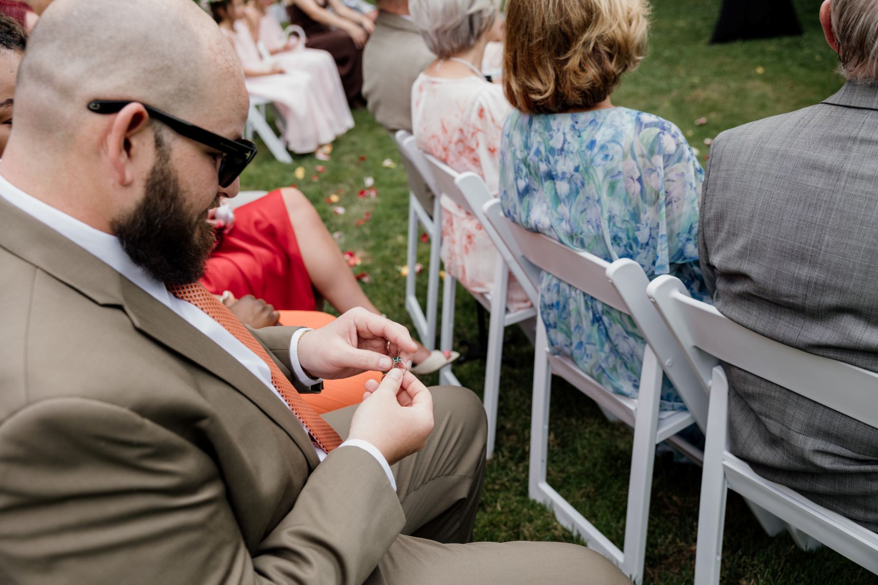 a man in a suit and sunglasses sitting in a white chair and holding a wedding ring surrounding him are other wedding guests 