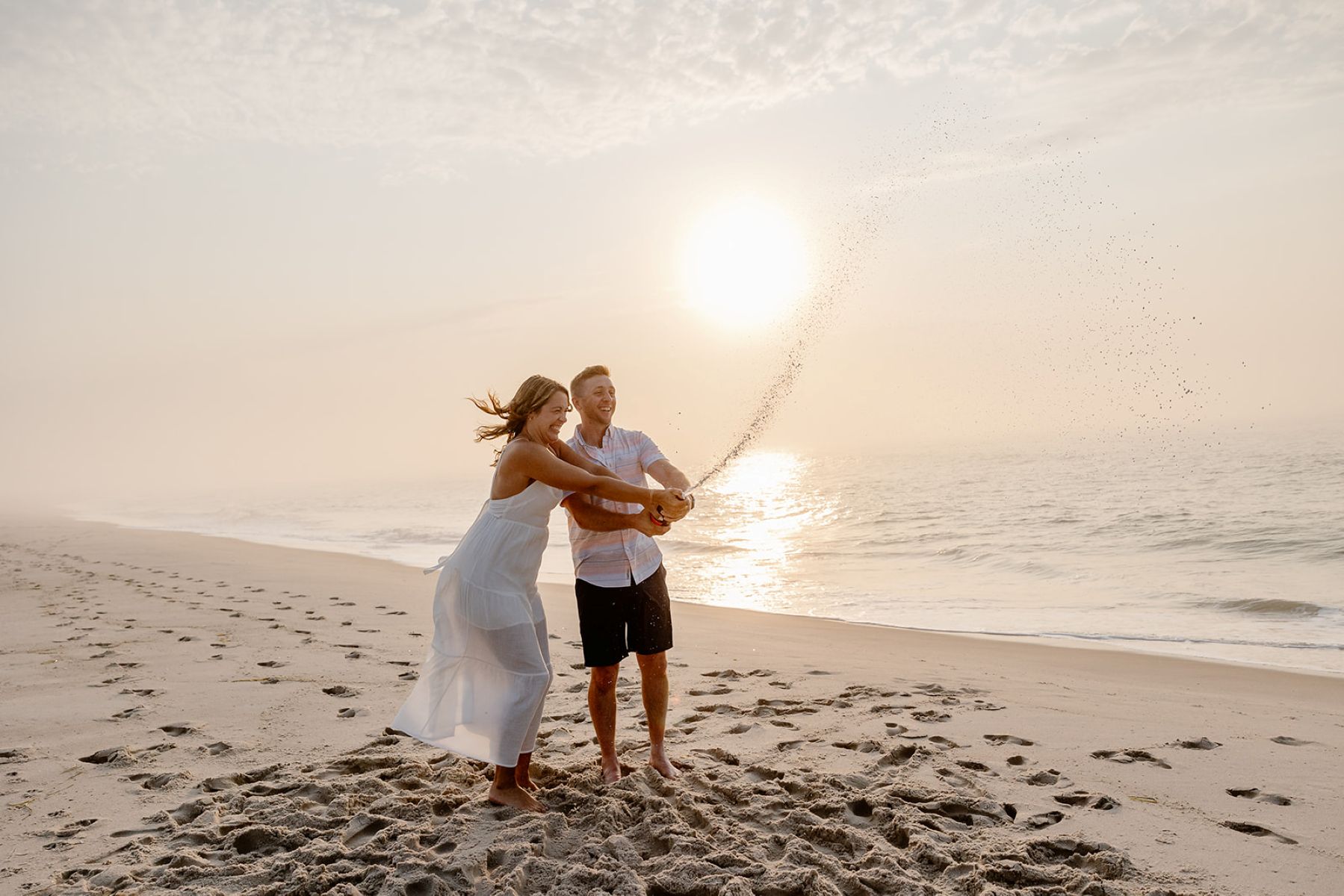 a couple on the sand next to the ocean popping a bottle of champagne together and laughing 