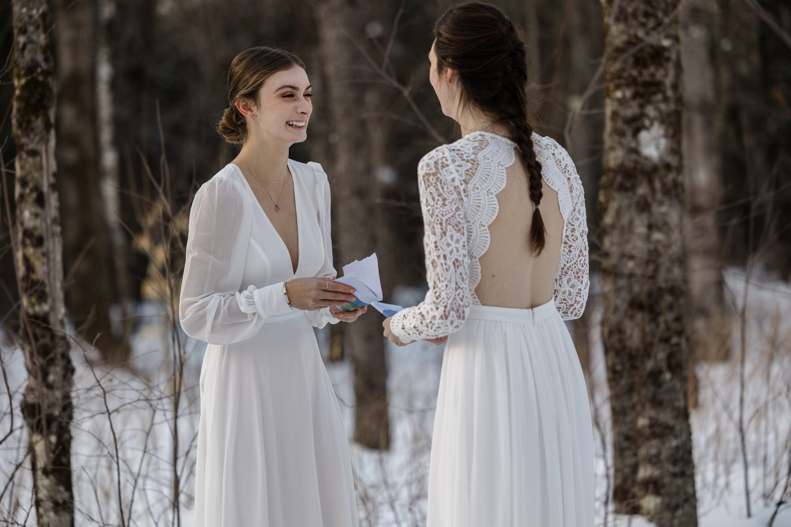 Two brides exchange vows in the snowy New Hampshire mountains. They are holding personal vow books, and smiling.