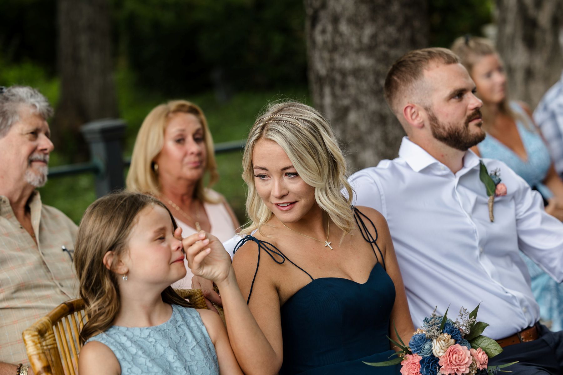 A yound girl is tearing up and a woman next to her is wiping a tear from her eye they are sitting with other wedding guests in wooden chairs 