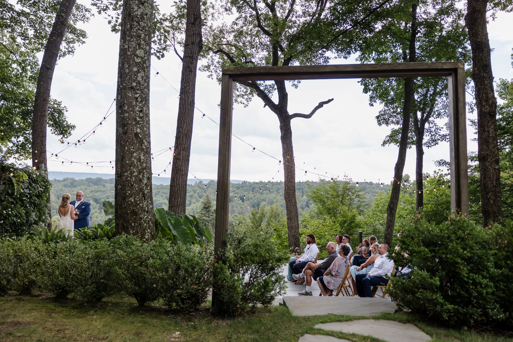 Wedding guests sitting and watching a wedding ceremony the couple is standing together in front of them underneath string lights and they are surrounded by plants and trees 