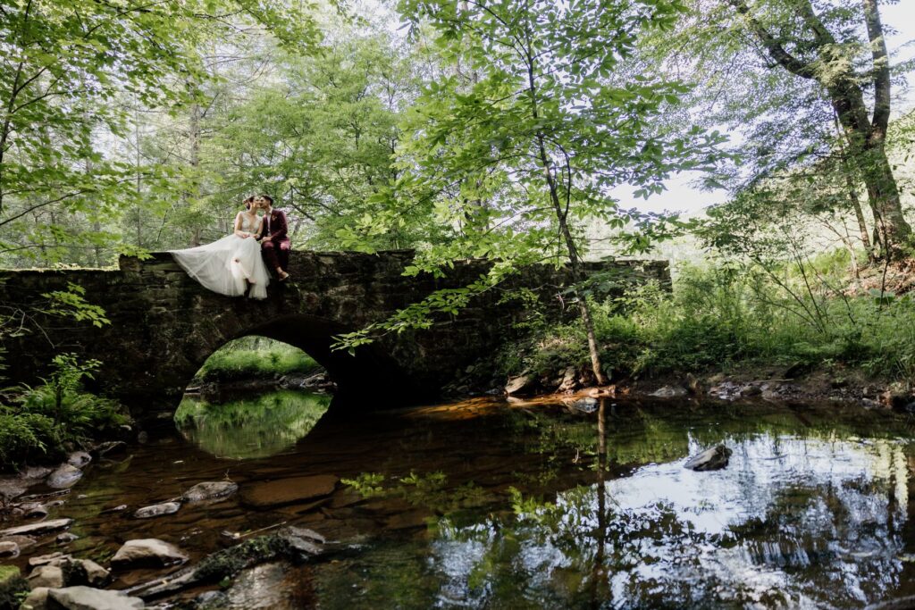 A bride and groom sit on an old stone bridge in the middle of a PA forest. 