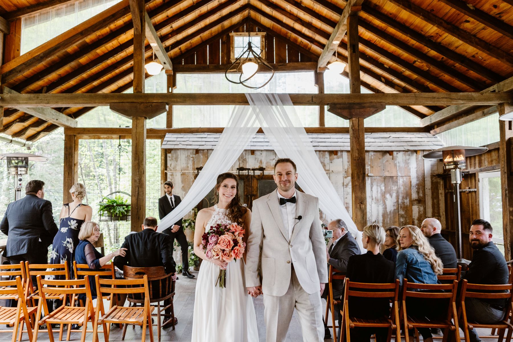 A couple walking together down the aisle smiling behind them their wedding guests are watching and sitting in wooden charits they are in a barn like structure the woman is wearing a white dress and holding a bouquet of flowers the man is wearing a tan suit